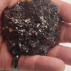 May include: A dark, crystalline mineral specimen with a rough, textured surface. The mineral has a deep brown color with reflective, shiny facets. The image shows the mineral held in a hand, highlighting its natural form and texture.