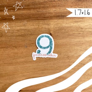 May include: A sticker with the number 9 in teal with a floral pattern, and the word "peacemaker" in pink script. The sticker measures 1.7 x 1.6 inches. The background is a wooden surface.
