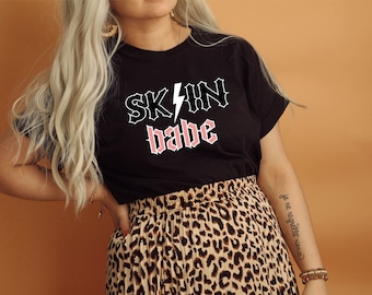May include: Black t-shirt with a graphic print that says "SK/N babe" in a gothic font with a lightning bolt. The model is wearing a leopard print skirt.