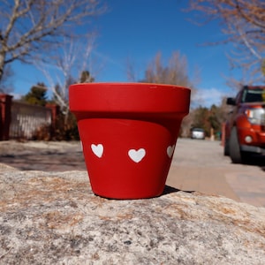 May include: Red terracotta flower pot with white heart designs. The pot is sitting on a gray rock.