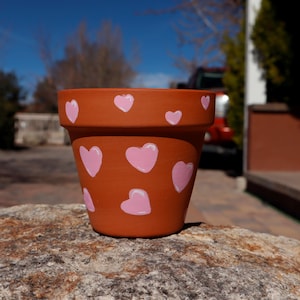 May include: A terracotta flower pot with a pink heart pattern. The pot is sitting on a rock.