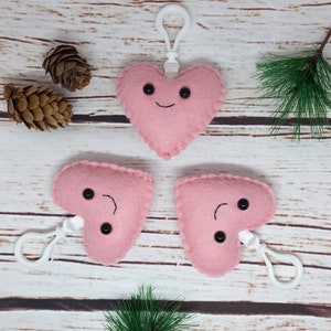 May include: Three pink felt heart-shaped keychains with smiling faces and black button eyes. Each keychain has a white clip. The hearts are on a light wood surface with pinecones and greenery.