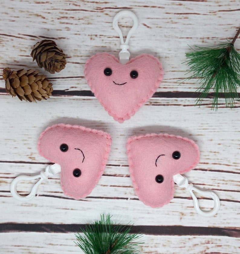 May include: Three pink felt heart-shaped keychains with smiling faces and black button eyes. Each keychain has a white clip. The hearts are on a light wood surface with pinecones and greenery.