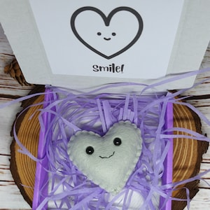 May include: A white felt heart with a smiling face rests in a purple box filled with shredded paper. The box is open, revealing a card with a heart graphic and the text "You make my heart Smile!". The box sits on a wooden slice.