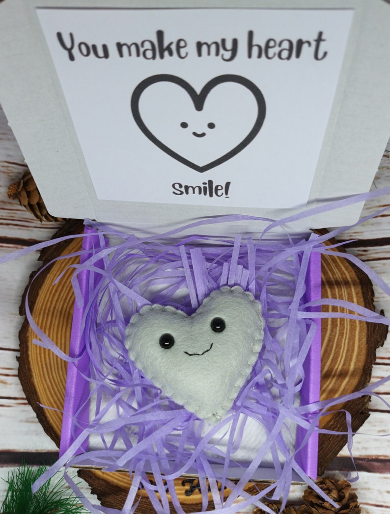 May include: A white felt heart with a smiling face rests in a purple box filled with shredded paper. The box is open, revealing a card with a heart graphic and the text "You make my heart Smile!". The box sits on a wooden slice.