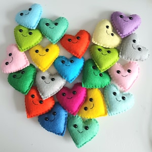 May include: A collection of colorful felt hearts arranged in a heart shape. Each heart is a different color, including pink, blue, green, yellow, orange, and gray, and has a smiling face.