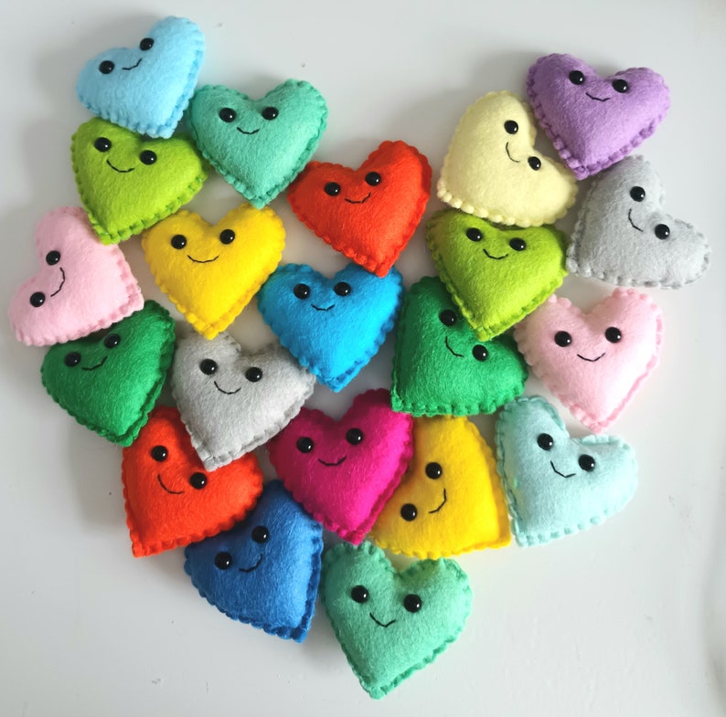 May include: A collection of colorful felt hearts arranged in a heart shape. Each heart is a different color, including pink, blue, green, yellow, orange, and gray, and has a smiling face.