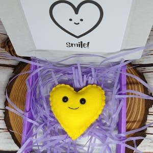 May include: A yellow felt heart with a smiling face rests in a box filled with purple paper shreds. The box contains a card with the text "You make my heart smile!" The box is on a wooden surface.