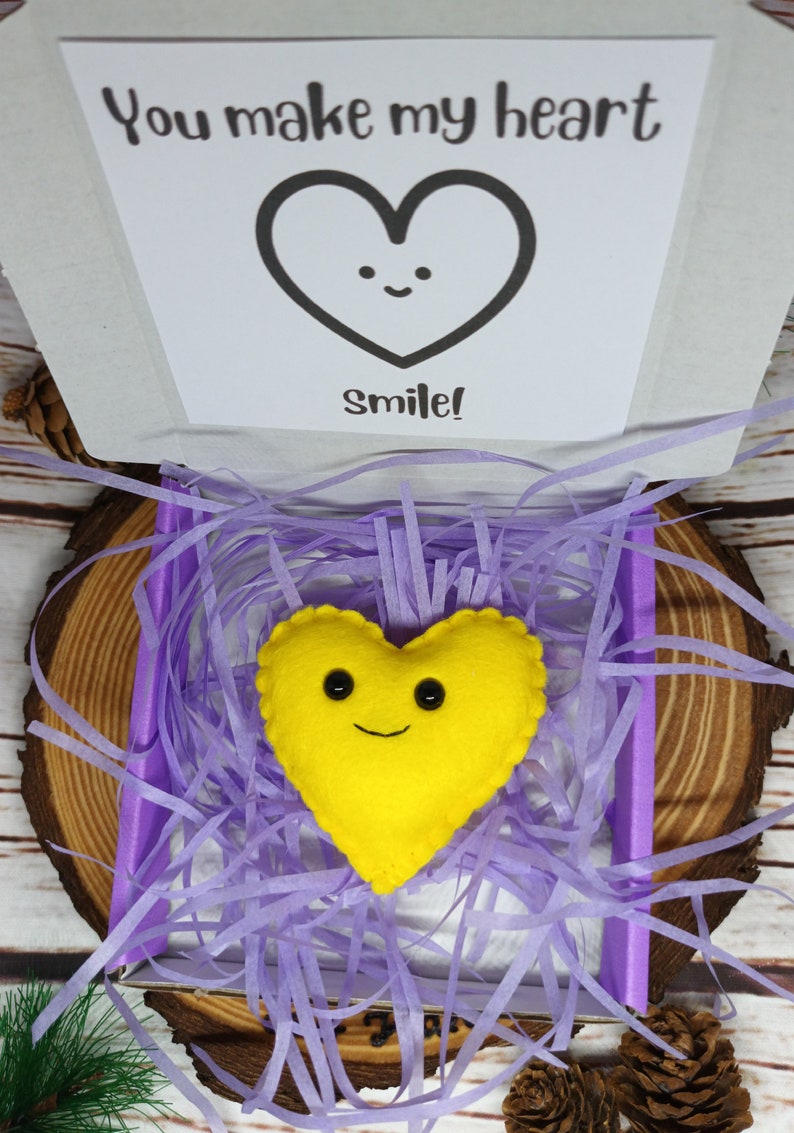 May include: A yellow felt heart with a smiling face rests in a box filled with purple paper shreds. The box contains a card with the text "You make my heart smile!" The box is on a wooden surface.