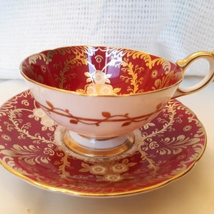 May include: A red and gold teacup and saucer set with a floral pattern. The cup and saucer are both decorated with a delicate floral design in gold.