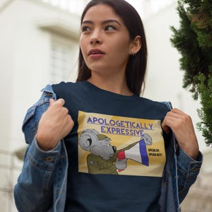 Puede incluir: Una camiseta azul marino con un koala de dibujos animados que lleva un uniforme militar y sostiene un megáfono. El texto de la camiseta dice "Apologetically Expressive" y "Speak Up, Speak Out!"