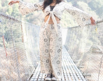 May include: A woman wearing a white crop top and matching pants with a celestial print. The top is tied at the waist and the pants are wide-legged. She is standing on a suspension bridge with a metal grate.
