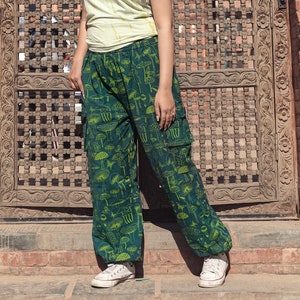 May include: A woman wearing a light yellow t-shirt and green pants with a mushroom print stands in front of a carved wooden door. The door is arched and has a lattice design. The woman is wearing white sneakers.