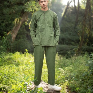 May include: A man wearing a green long-sleeved shirt and matching pants stands in a forest setting. The shirt has a button-up collar and a single button closure. The man is looking directly at the camera.