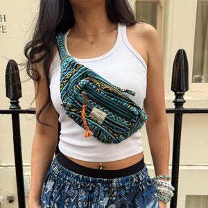 May include: A teal, black, and yellow woven fanny pack featuring a geometric pattern, a front zipper with a multi-colored braided pull, and a rectangular fabric label with the text "Himalayan Handmades" worn across the chest over a white tank top.
