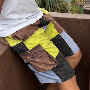 May include: A person wearing a white t-shirt and patchwork shorts with brown, blue, black, and yellow fabric. The shorts have a large cargo pocket on the right side.