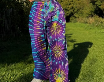 May include: Loose-fitting pants featuring a vibrant tie-dye pattern in shades of purple, blue, yellow, and pink. The garment includes an elastic waistband with a drawstring tie and a distinct sunburst design on the legs, paired with white sneakers.