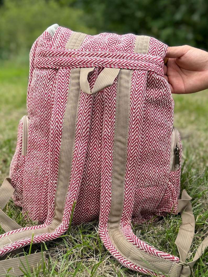 Op de afbeelding: Een rode en witte rugzak met visgraatpatroon en bruine bandjes en accenten. De rugzak heeft twee zijzakken met ritsen en verstelbare schouderbanden. De tas is gemaakt van een geweven stof.