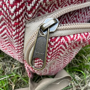 Op de afbeelding: Close-up van een rood en wit patroon rugzak met een beige rits en riem. De ritstrekker is donker metaalkleurig. De rugzak is gemaakt van een geweven stof.