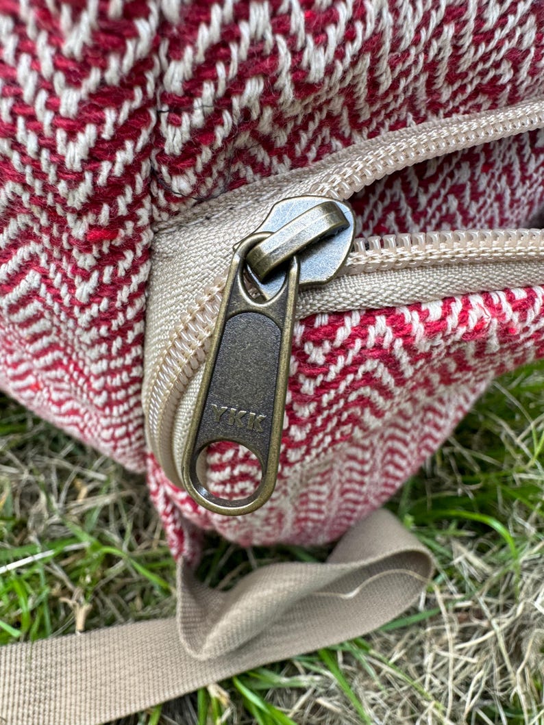 Op de afbeelding: Close-up van een rood en wit patroon rugzak met een beige rits en riem. De ritstrekker is donker metaalkleurig. De rugzak is gemaakt van een geweven stof.