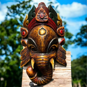 May include: A wooden carved mask of the Hindu deity Ganesha, painted in shades of brown, red, and gold. The mask features a detailed elephant head with a trunk curled up, large ears, and a crown with intricate carvings.