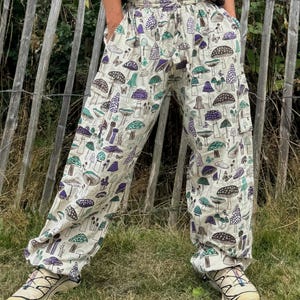 May include: White trousers with a mushroom print. The trousers have an elastic waistband and a drawstring. The mushrooms are in shades of purple, green, and brown.