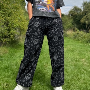 May include: Black cargo trousers featuring a celestial print of suns, moons, and stars. The trousers have multiple pockets and a relaxed fit. The person is wearing a dark grey t-shirt with a sun graphic.