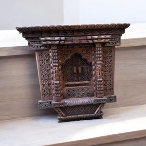 May include: A dark brown wooden wall decoration with intricate carvings. The decoration features a window-like design with a smaller window inside. The decoration is made of wood and has a rustic look.