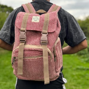 Op de afbeelding: Rode en beige rugzak met visgraatpatroon. De rugzak heeft verstelbare banden, een bovenklep met twee gespsluitingen en een voorvak met ritssluiting. De riemen en gespen zijn beige. De rugzak is gemaakt van geweven materiaal.