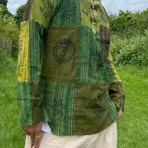 May include: A green and yellow patchwork long-sleeved shirt with various printed designs, including a sun, a flower, and a mushroom. The shirt has a wooden button closure at the neck.