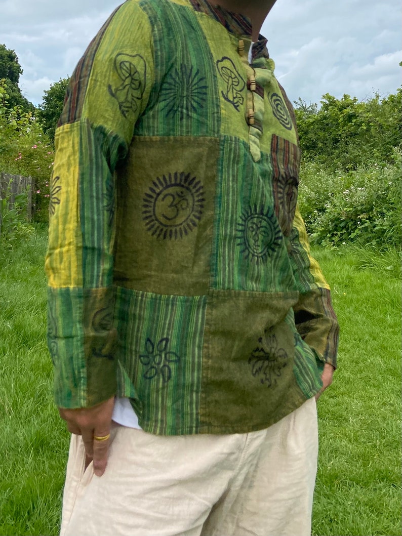May include: A green and yellow patchwork long-sleeved shirt with various printed designs, including a sun, a flower, and a mushroom. The shirt has a wooden button closure at the neck.