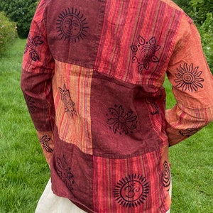 May include: A long-sleeved shirt with a patchwork design featuring various symbols and patterns. The shirt is made of a red and orange striped fabric with black printed details. The symbols include suns, flowers, and a symbol that looks like an Om.