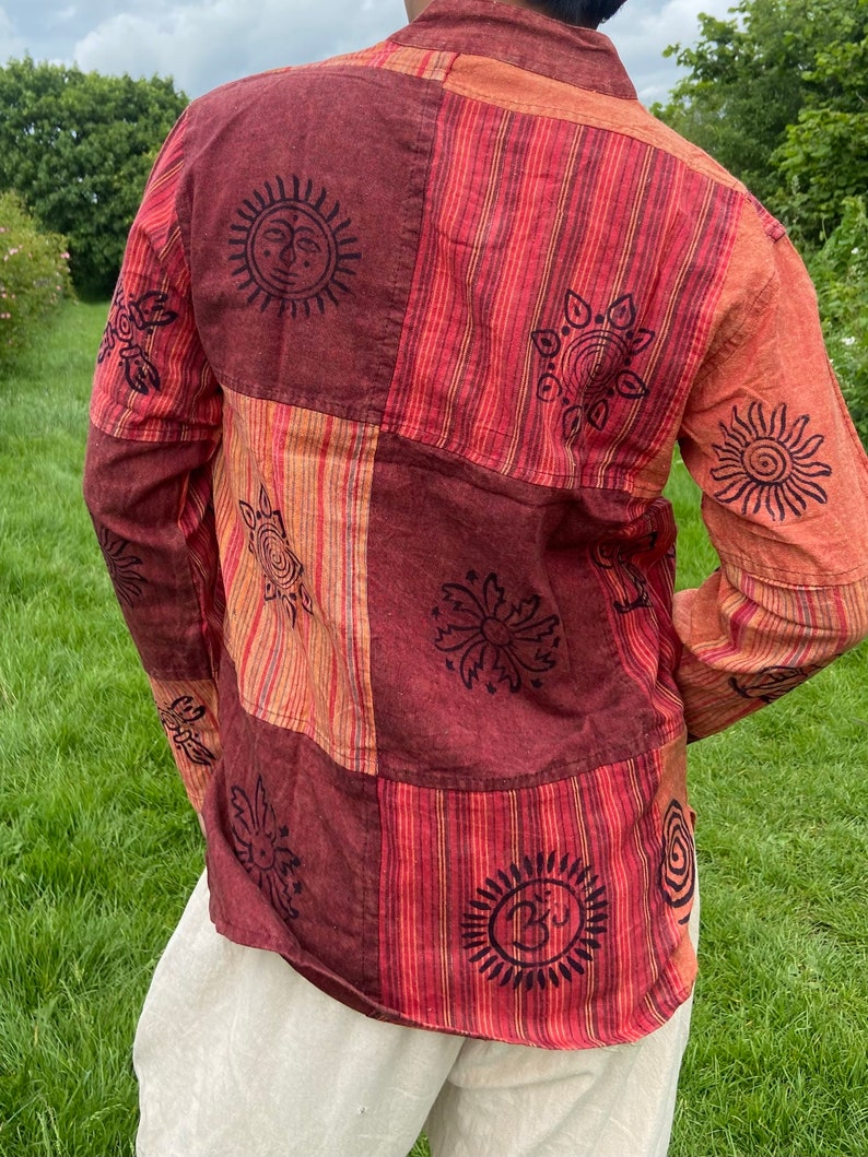 May include: A long-sleeved shirt with a patchwork design featuring various symbols and patterns. The shirt is made of a red and orange striped fabric with black printed details. The symbols include suns, flowers, and a symbol that looks like an Om.