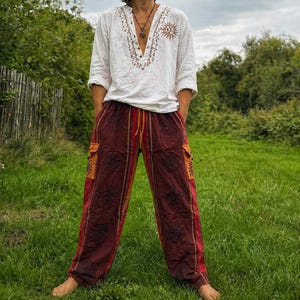 May include: A white long-sleeved shirt with embroidered detailing and a sun motif, worn with maroon trousers featuring floral patterns and orange accents. The trousers have a drawstring waist and pockets. The outfit is worn outdoors.