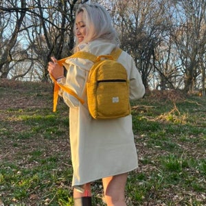 Peut inclure: Sac à dos jaune moutarde avec une poche avant et une fermeture éclair noire. Le sac à dos a des bretelles réglables et une petite étiquette sur le devant. La personne porte un manteau crème dans une zone boisée.