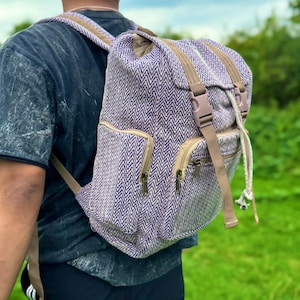 Op de afbeelding: Een paarse en witte rugzak met visgraatpatroon en bruine bandjes en accenten. De rugzak heeft twee voorzakken met rits, een trekkoordsluiting en een bovenklep met een gespsluiting. De tas wordt over de schouder gedragen.