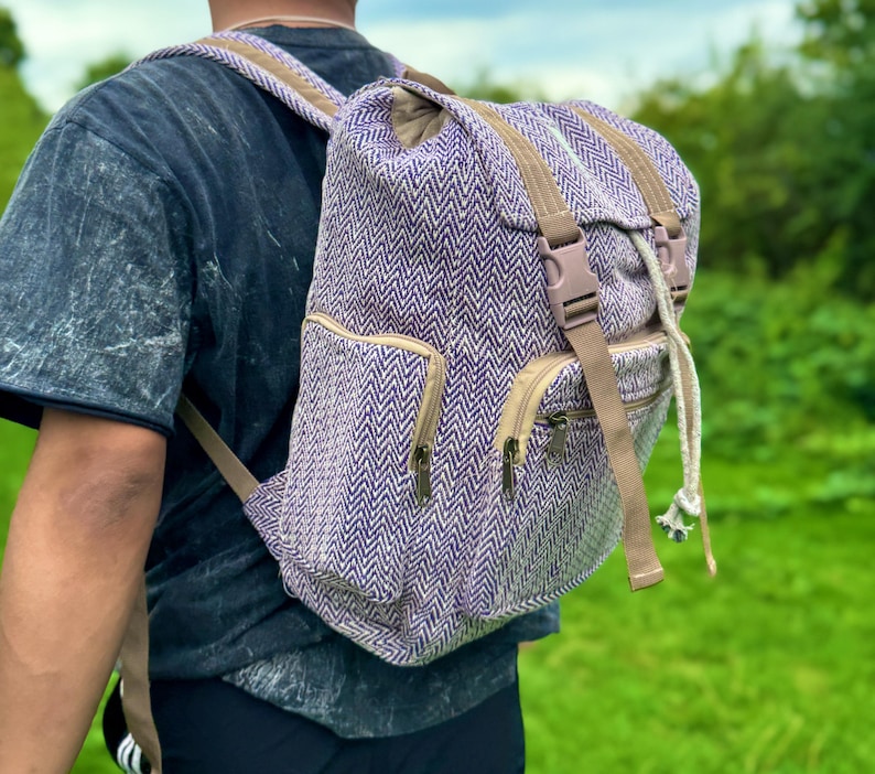 Op de afbeelding: Een paarse en witte rugzak met visgraatpatroon en bruine bandjes en accenten. De rugzak heeft twee voorzakken met rits, een trekkoordsluiting en een bovenklep met een gespsluiting. De tas wordt over de schouder gedragen.