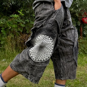 May include: Acid-washed black cargo shorts featuring a large white circular geometric mandala-style print on the side pocket. The shorts have a loose, wide-leg fit with visible frayed edges, multiple cargo pockets, and a distressed, mottled fabric texture.