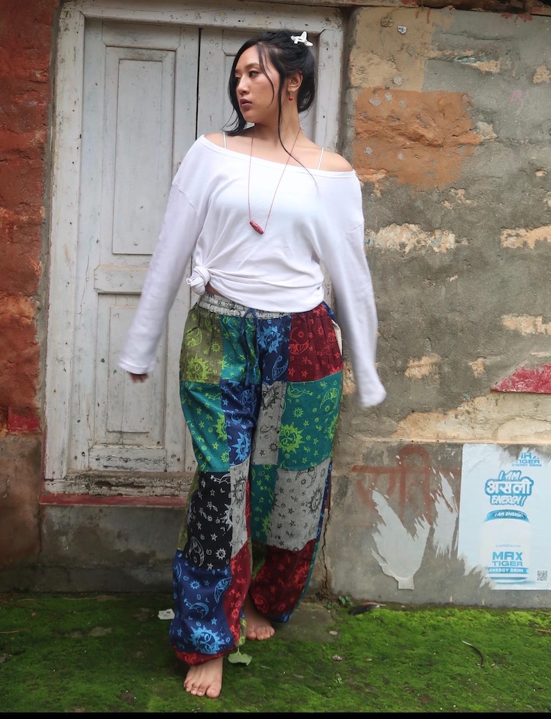 Puede incluir: Una camisa blanca de manga larga con la cintura anudada y pantalones de patchwork holgados con un patr&oacute;n multicolor de soles, lunas y estrellas en tonos azul, verde, rojo y negro con una cintura el&aacute;stica con cord&oacute;n.