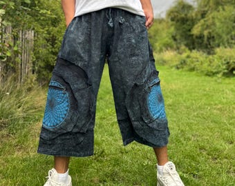 May include: Dark charcoal gray cropped pants with a distressed, acid-wash texture. The pants feature a drawstring waist and large, circular side pockets with bright blue geometric mandala-style prints and black zipper closures on each leg.