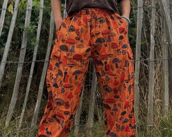 Peut inclure: Pantalon orange orné d'un motif répétitif de champignons rouges, bleus et noirs. Le pantalon a une coupe ample et des poches. La personne porte un t-shirt marron avec un petit motif.