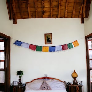 May include: A bedroom scene featuring a bed with a decorative headboard, flanked by wooden nightstands. A string of colorful prayer flags hangs across the wall, with a framed picture above the bed. The ceiling is wood-paneled.