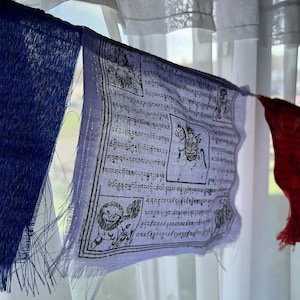 May include: A string of colorful prayer flags hanging in front of a window with sheer white curtains. The flags are rectangular, with a variety of colors including blue, red, and white. Each flag features intricate black and white designs and text.