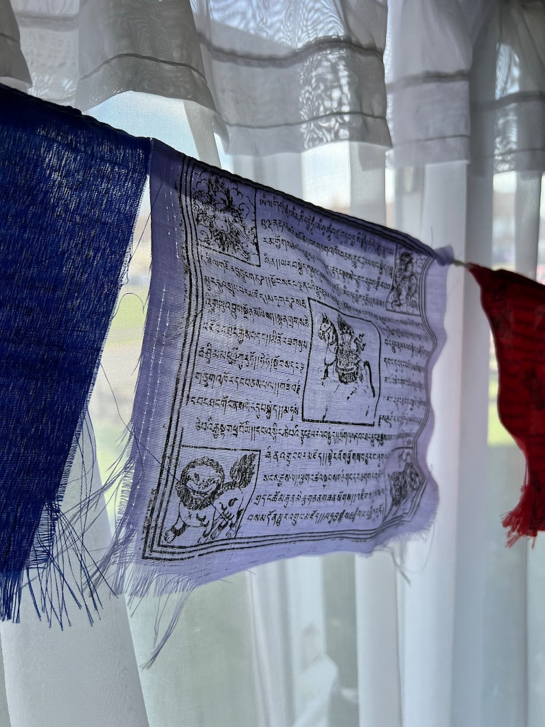 May include: A string of colorful prayer flags hanging in front of a window with sheer white curtains. The flags are rectangular, with a variety of colors including blue, red, and white. Each flag features intricate black and white designs and text.