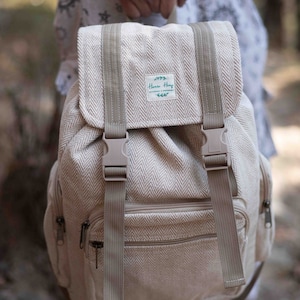 Może przedstawiać: Beżowy plecak z wzorem w jodełkę, z regulowanymi paskami i klapą z dwoma klamrami. Torba posiada przednią kieszeń na zamek i małą etykietę z napisem "Hanno Hemp".