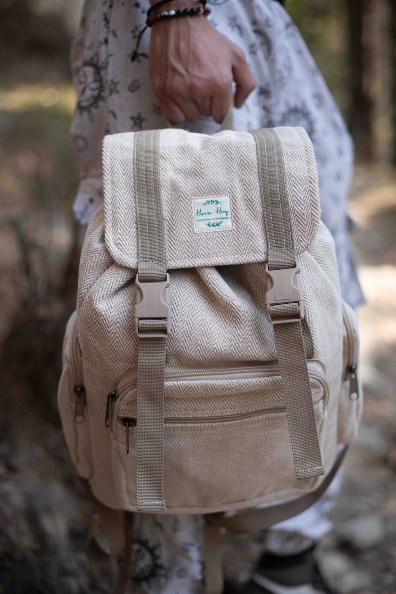 Op de afbeelding: Beige rugzak met een visgraatpatroon, verstelbare bandjes en een klepsluiting met twee gespen. De tas heeft een voorvak met rits en een klein label met de tekst "Hanno Hemp".