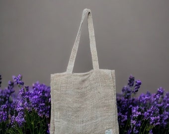 May include: A beige tote bag with long handles, made of a woven fabric. The bag is rectangular with a small label on the front. Purple flowers are in the foreground, and a gray wall is in the background.