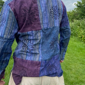 May include: A long-sleeved patchwork shirt in blue and purple hues. The shirt features a repeating sun design with a face in the centre.