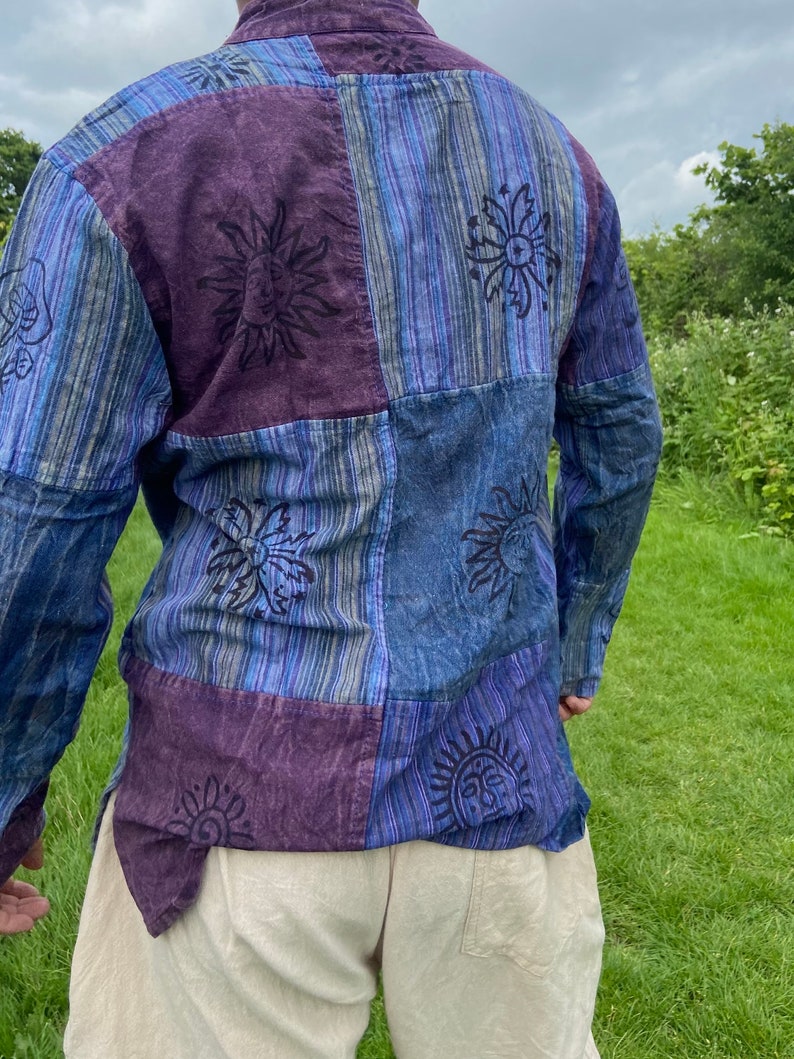 May include: A long-sleeved patchwork shirt in blue and purple hues. The shirt features a repeating sun design with a face in the centre.
