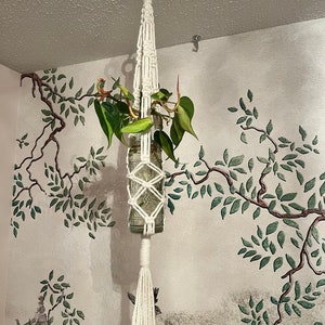 May include: White macrame plant hanger with a glass bottle and a green plant hanging from the ceiling against a white wall with a green tree design.
